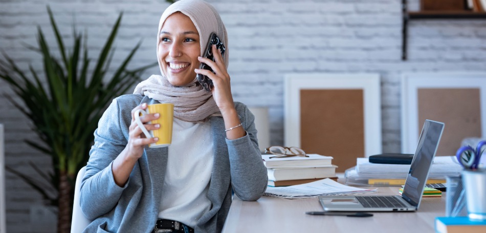 Person sitting at a desk holding a phone and a yellow mug with an open laptop and office supplies in the background, representing mobile banking